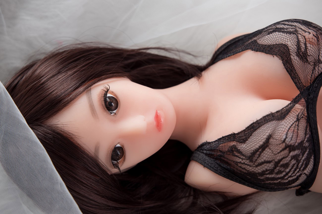 Realistic love doll with long dark hair and black lace lingerie lying on bed.